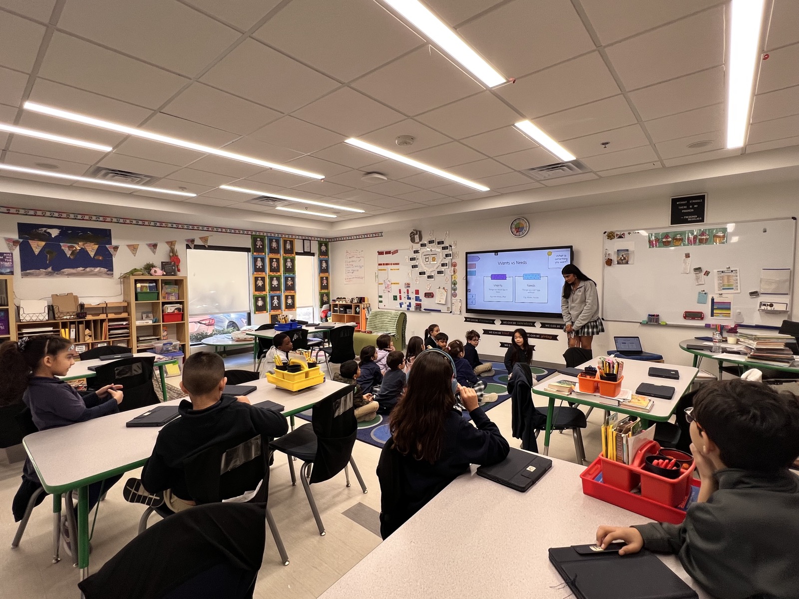 A BizBuddies volunteer standing at the front of an elementary classroom, teaching a wants-versus-needs lesson to seated students